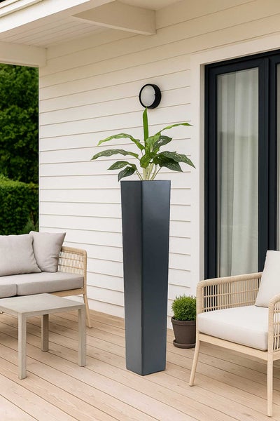 Moderner hoher, quadratischer Pflanzkübel in Anthrazit auf einer hellen Holzterrasse mit Lounge-Möbeln vor einer Fassade.