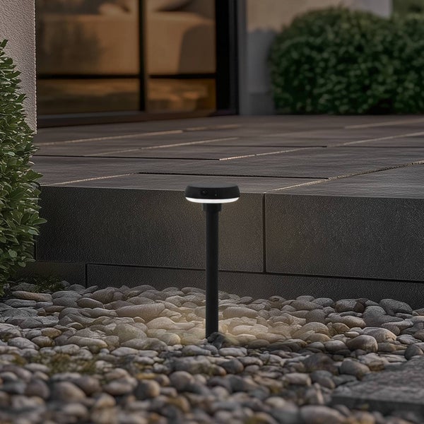 Eine schwarze LED-Außenleuchte in einem Kiesbeet vor einer modernen Steinterrasse bei Dämmerung, die warmes Licht auf die Steine wirft.