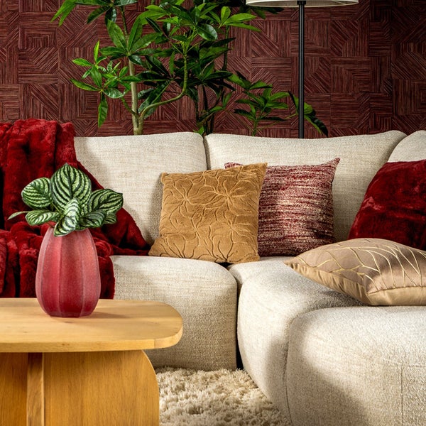 Wohnzimmer mit beigem Polstersofa, Dekokissen, roter Kuscheldecke, Holztisch mit Zimmerpflanze und strukturierter Tapete mit geometrischem Muster.
