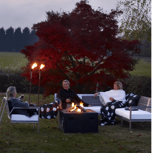 Gas-Feuerstelle im Garten mit Aluminium-Sitzgruppe, brennendem Feuer, Gartenfackeln und Personen in herbstlicher Atmosphäre.
