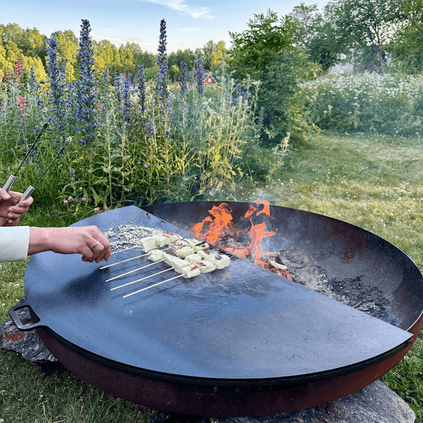 Eine runde Feuerschale aus Metall mit einer halbkreisförmigen Grillplatte in einem Garten. Eine Person grillt Spieße über einem Holzfeuer.