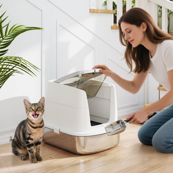 Eine Frau reinigt eine moderne Katzentoilette mit Edelstahlschale und weißer Haube, während eine Katze daneben sitzt.