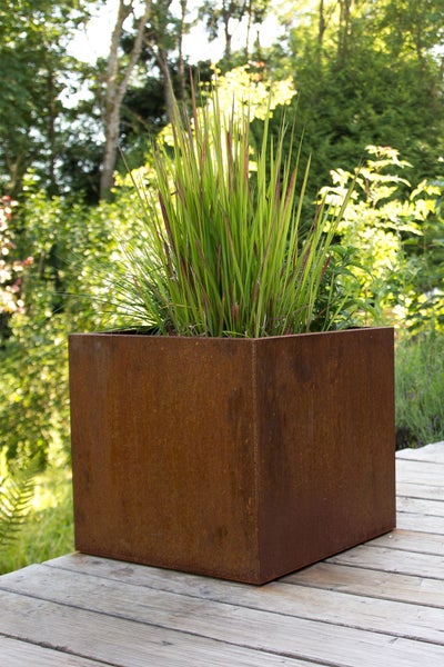 Quadratischer Pflanzkübel aus Cortenstahl mit Rostoptik auf einer Holzterrasse, bepflanzt mit hohem Ziergras im Garten.