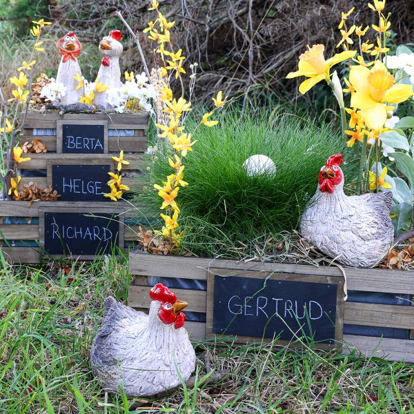 Osterdekoration mit Hühnerfiguren aus Keramik in Holzstiegen mit beschrifteten Tafeln und Frühlingsblumen im Garten.
