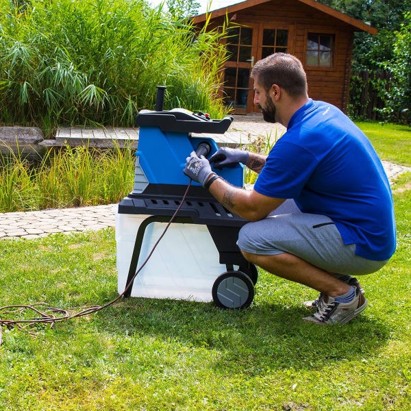 Mann schließt Kabel an blauen Gartenhäcksler mit Auffangbehälter an. Im Hintergrund ein Garten mit Teich und Holzhütte.