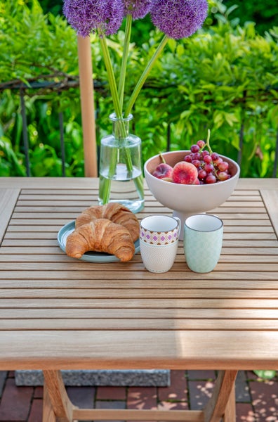 Gartentisch aus Holz auf einer Terrasse mit gedecktem Frühstück, belegt mit Croissants, Obstschale, Tassen und einer Blumenvase.