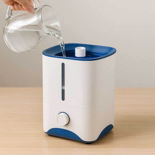 Befüllen eines Luftbefeuchters mit Wasser aus einem Glaskrug von oben auf einem Holztisch.