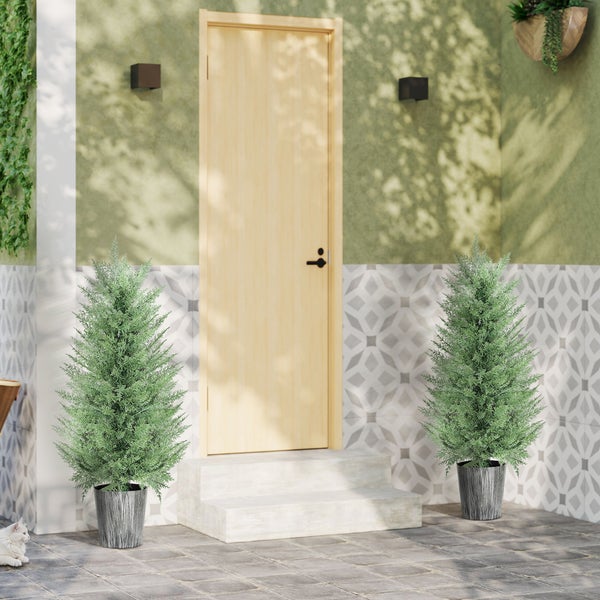 Hauseingang mit heller Holztür, Steinstufen und zwei künstlichen Zypressen in Töpfen auf Steinboden vor gemusterter Wand.