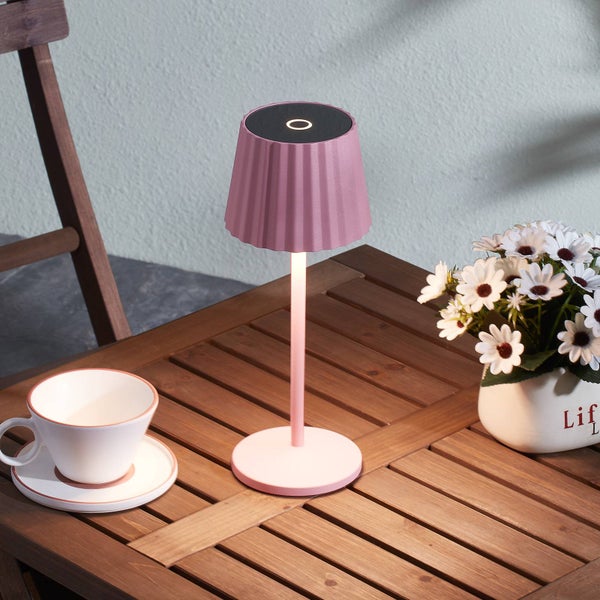 Moderne Tischleuchte mit plissiertem Schirm auf einem Holztisch neben einer Tasse und Blumen in einer hellen Außenumgebung.