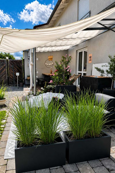 Gemütliche Terrasse mit einem Sonnensegel, schwarzen Pflanzkübeln mit Ziergras, Loungemöbeln und blühenden Pflanzen für eine wohnliche Atmosphäre.