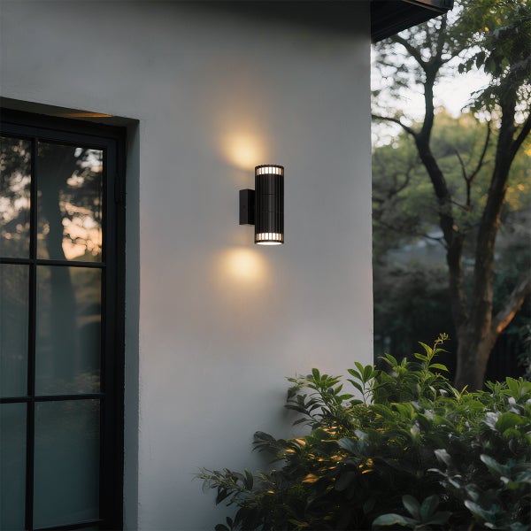 Zylindrische schwarze Außenwandleuchte an weißer Fassade mit Licht nach oben und unten, neben Fenster und grünen Gartenpflanzen.