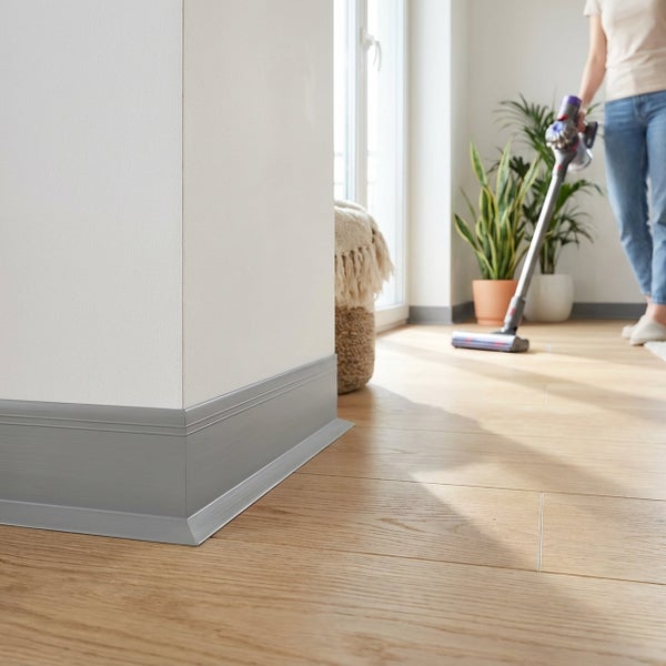 Moderne Fußleiste in einem Wohnraum mit Holzboden, während eine Person mit einem Akku Staubsauger den Raum reinigt.