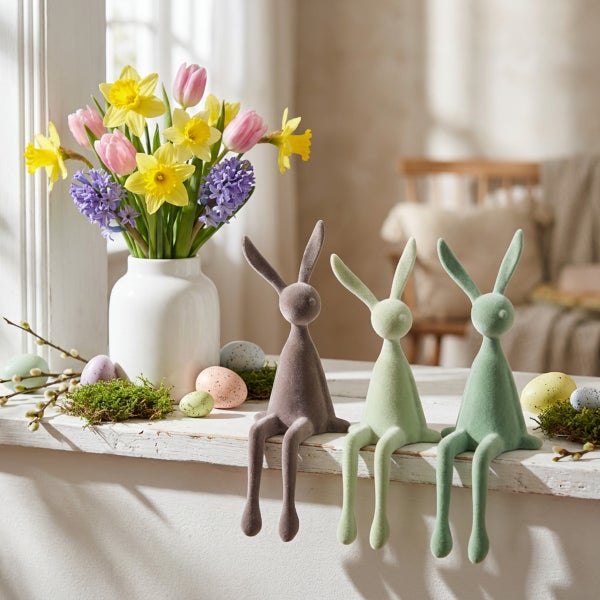 Osterdekoration mit drei Osterhasen Figuren aus beflocktem Material, einer Blumenvase mit Frühlingsblumen und dekorativen Ostereiern auf Fensterbank.