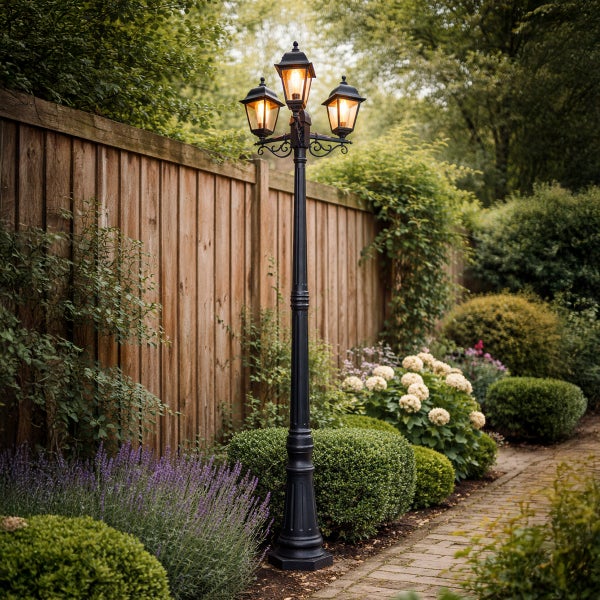 Dreiflammige Gartenlaterne aus schwarzem Metall vor einem Holzzaun in einem bepflanzten Garten mit gepflastertem Weg.