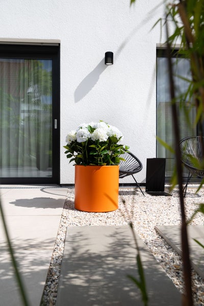 Oranger zylindrischer Pflanzkübel mit weißen Hortensien auf einer modernen Terrasse mit Kies, Gehwegplatten und schwarzen Gartenstühlen.