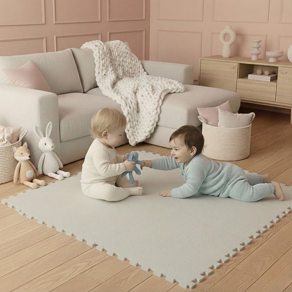 Zwei Babys spielen auf einer quadratischen Puzzlematte aus Schaumstoff in einem hellen Raum mit Sofa und Holzfußboden.