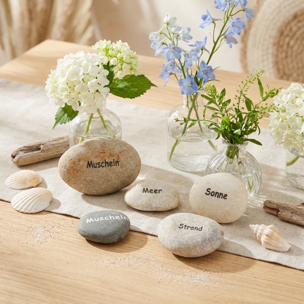 Dekorative Kieselsteine mit Aufschrift Meeresthema neben kleinen Glasvasen mit Blumen und Muscheln auf einem Tisch.