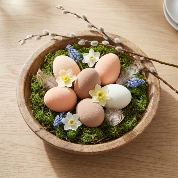 Osterschale aus Holz gefüllt mit Eiern, Moos, Narzissen, Traubenhyazinthen und Weidenkätzchenzweigen als frühlingshafte Tischdekoration.