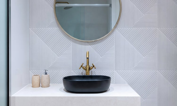 Modernes Badezimmer mit schwarzem Aufsatzwaschbecken, messingfarbener Armatur und hellen Wandfliesen mit geometrischer Struktur.