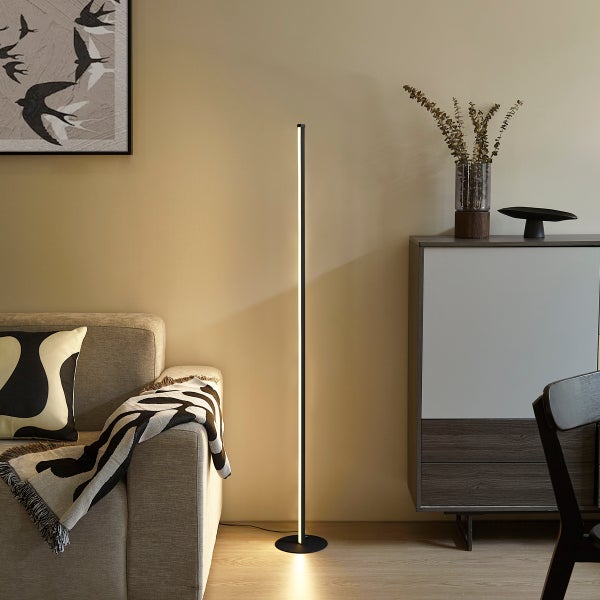 Moderne minimalistische LED-Stehleuchte in Stabform neben einem Sofa im Wohnzimmer, schwarzes Gehäuse mit warmweißem Licht.