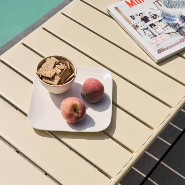 Hellbeiger Gartentisch aus Metall mit Lattenstruktur, am Pool mit Pfirsichen, Schale mit Crackern, Magazin und Glas Wasser.