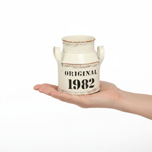 Kleine cremefarbene Deko-Milchkanne aus Metall im Vintage-Stil mit Aufdruck Original 1982, in einer Hand präsentiert.