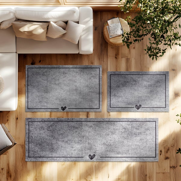 Drei graue Teppiche in verschiedenen Größen mit Herzmotiv und Rand auf hellem Holzboden in einem hellen Wohnzimmer mit Sofa und Pflanze.