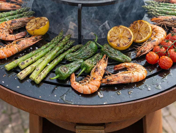 Garnelen, grüner Spargel, Paprika, Zitronen und Tomaten grillen auf einer runden Feuerstelle mit Grillring aus Metall.
