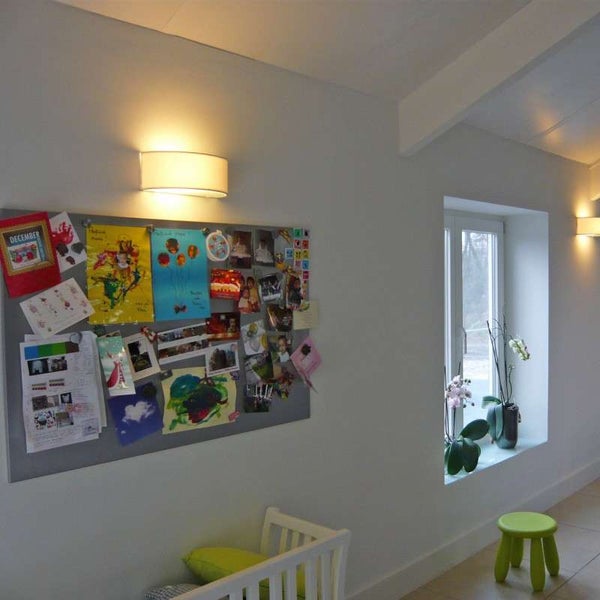 Wandleuchte mit warmem Licht über einer Magnetwand mit Fotos in einem hellen, modern eingerichteten Raum mit Fenster.