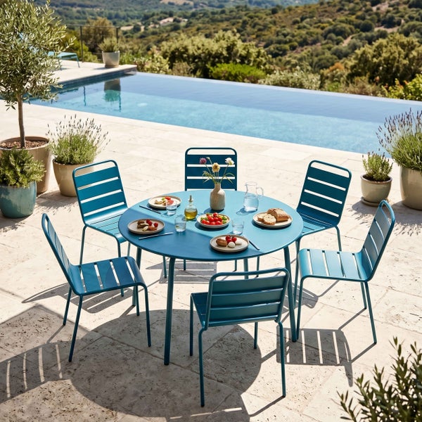 Helle Terrasse mit blauem Gartenmöbel-Set aus Metall, runder Tisch und sechs Lattenstühle, gedeckt mit Speisen, mit Blick auf Pool und Landschaft.