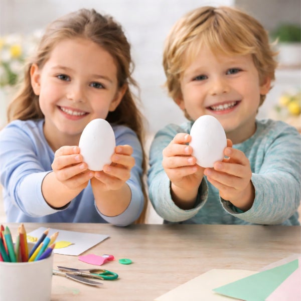 Zwei Kinder präsentieren Ostereier an einem Basteltisch mit Scheren, Stiften und Papier für ein österliches Bastelprojekt.