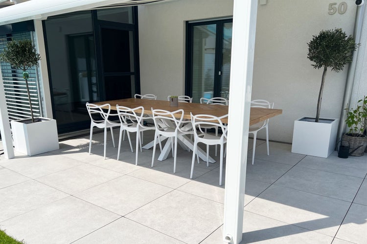 Moderne Terrasse mit großem Holztisch, weißen Stühlen mit Ziermuster und Olivenbäumen in Kübeln auf hellen Bodenfliesen.