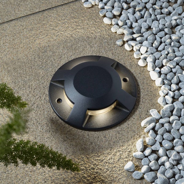 Runder schwarzer Bodeneinbaustrahler auf Steinplatten neben Kieselsteinen mit seitlichem warmem Licht.