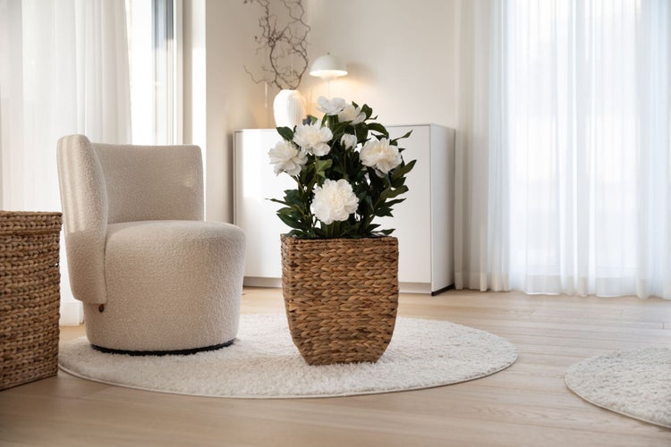 Hell eingerichtetes Wohnzimmer mit einem geflochtenen Pflanzkorb voller weißer Kunstpfingstrosen neben einem beigen Sessel aus Bouclé-Stoff.