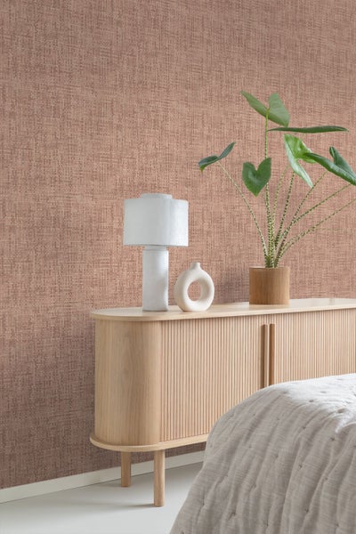 Vliestapete mit Leinenstruktur in einem hellen Schlafzimmer mit Sideboard aus Holz, weißer Tischleuchte und Zimmerpflanze.