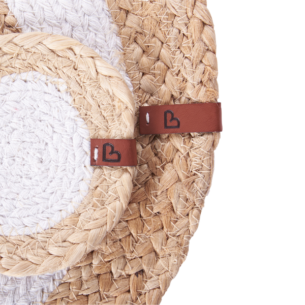 Nahaufnahme von zwei runden, geflochtenen Tischsets aus Jute und weißem Stoff, jeweils mit einem braunen Lederetikett mit Logo.