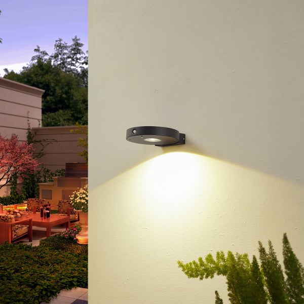 Schwarze, runde LED-Außenwandleuchte mit Bewegungsmelder an einer Fassade, die einen Terrassenbereich bei Dämmerung beleuchtet.