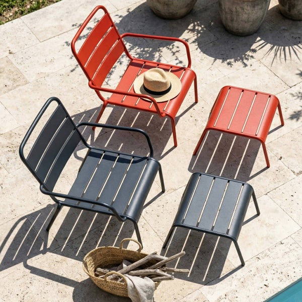 Moderne Gartenlounge auf einer Steinterrasse mit Gartenstühlen aus Metalllatten, passenden Fußhockern, einem geflochtenen Korb und einem Strohhut.