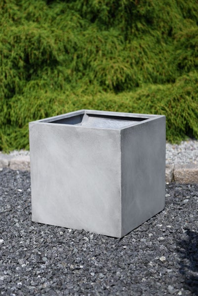 Würfelförmiger grauer Pflanzkübel in Betonoptik auf dunklem Kies im Garten vor einer grünen Hecke.
