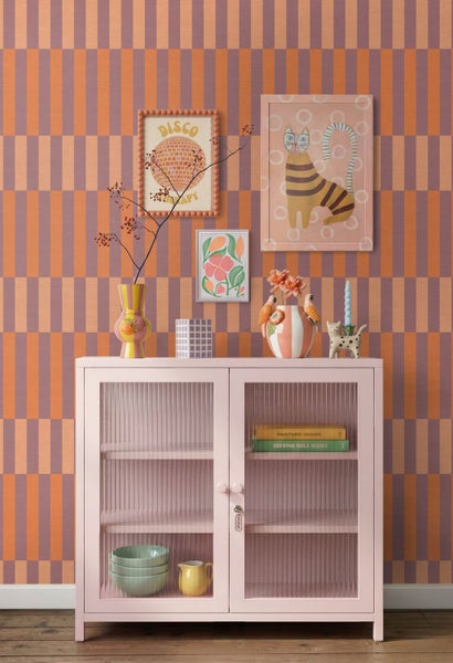 Ein hell eingerichtetes Zimmer mit einem rosa Metallschrank mit Riffelglastüren vor einer geometrisch gemusterten Tapete und Wanddekoration.