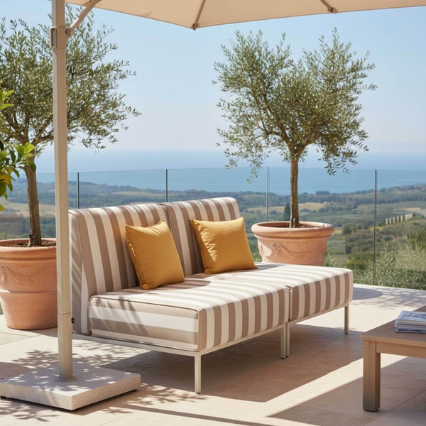 Gestreifte Gartenlounge mit gelben Kissen unter einem Sonnenschirm auf einer hellen Terrasse mit Olivenbäumen und Meerblick.