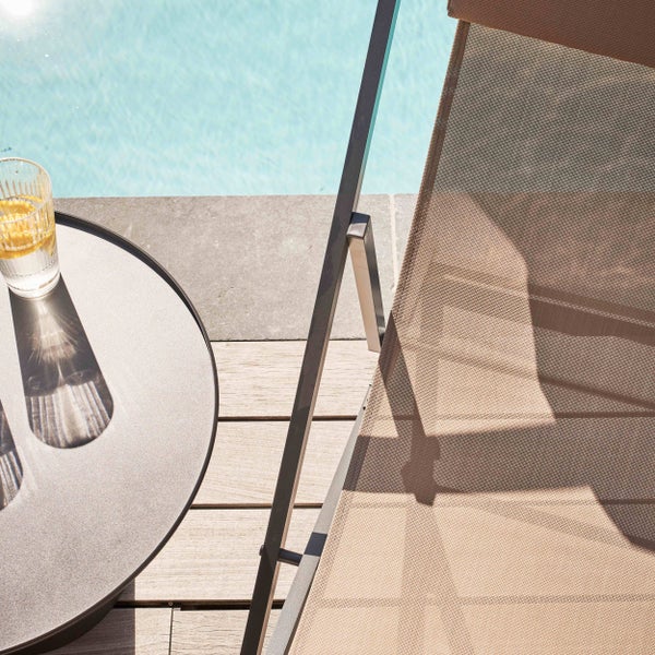 Sonnenliege mit Netzbespannung und Beistelltisch auf einem Holzdeck neben einem Pool.