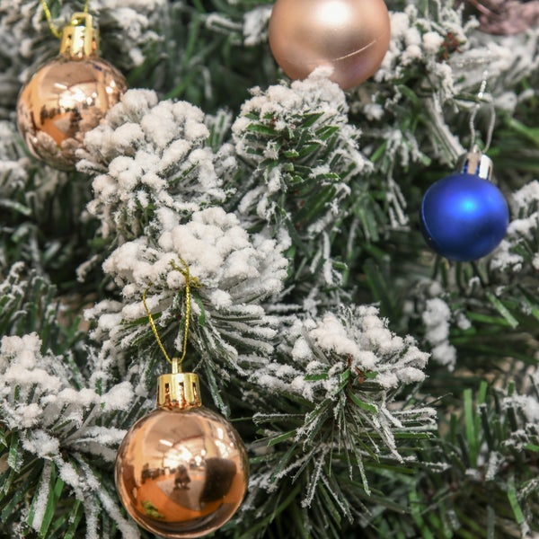 Nahaufnahme eines verschneiten künstlichen Weihnachtsbaums mit goldenen roségoldenen und blauen Christbaumkugeln.