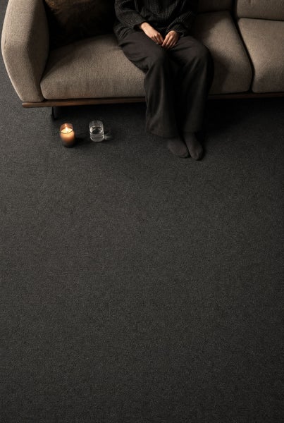 Dunkler, strukturierter Teppichboden in einem Wohnzimmer mit Sofa, sitzender Person und einer brennenden Kerze.