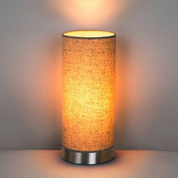 Zylinderförmige Tischleuchte mit grauem Textilschirm und silberfarbenem Metallfuß, eingeschaltet mit warmem Licht.