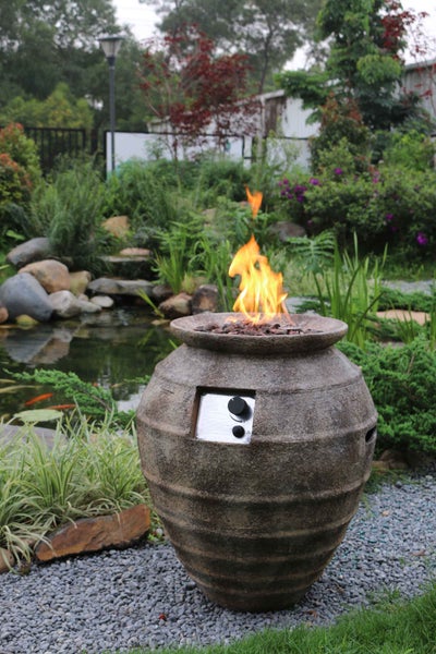 Gas-Feuerstelle in Form einer strukturierten Steinvase mit offener Flamme in einer Gartenlandschaft mit Teich und Kies.