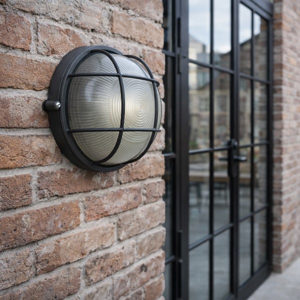 Runde Außenwandleuchte im Industrie-Stil mit schwarzem Metallschutzgitter und geripptem Glasdiffusor an einer Backsteinwand.