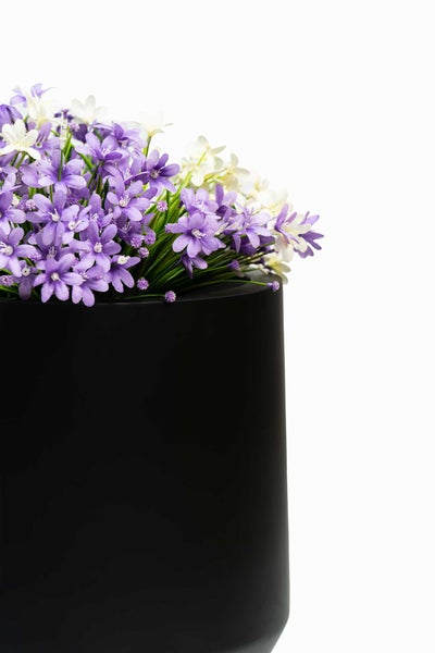 Künstliche lila und weiße Blumen in einem schwarzen zylindrischen Blumentopf.