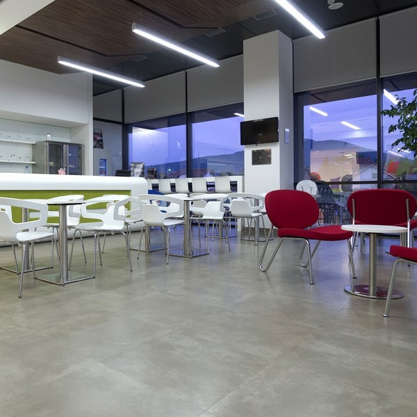 Moderner, weitläufiger Aufenthaltsbereich mit grauen Bodenfliesen, weißen und roten Kunststoffstühlen, Metalltischen und Glasfronten.