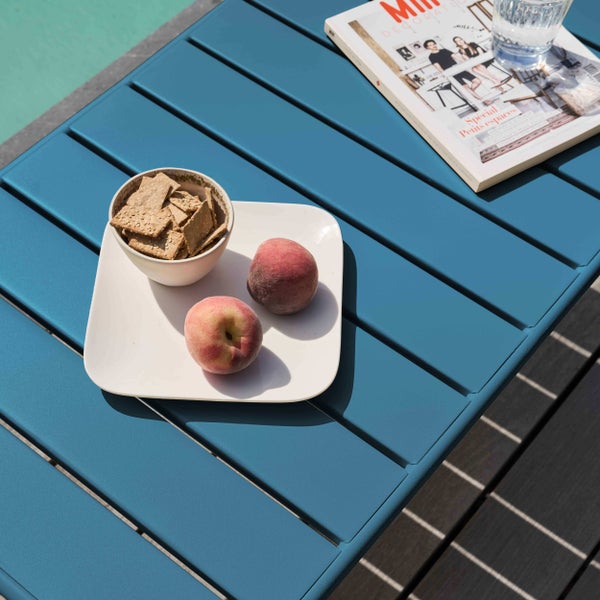 Blauer Gartenbeistelltisch aus Metall mit Lattenoberfläche, darauf ein Teller mit Pfirsichen, eine Schale Cracker und eine Zeitschrift.